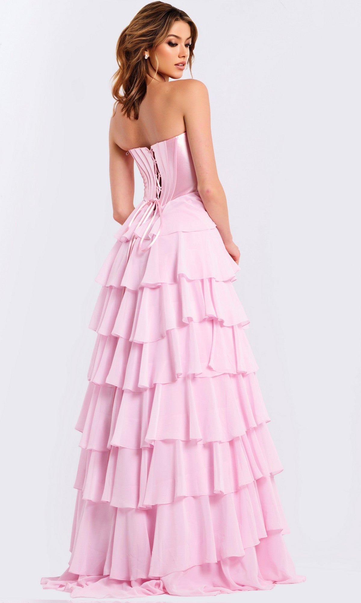 A woman wearing the Jovani 50234 long prom dress with a strapless neckline, chiffon ruffle skirt, and lace-up back stands facing away from the camera.